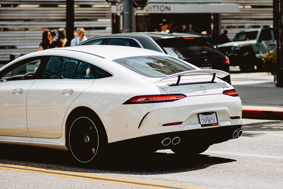 A sleek white luxury car cruising in an urban setting with people in the background on a sunny day